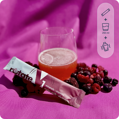 Rotate V1 Forest Fruit Stick vor einem Glas des zubereiteten Beerengetränks mit Zitronenscheibe und Eis, umgeben von frischen Beeren auf pinkfarbenem Stoff