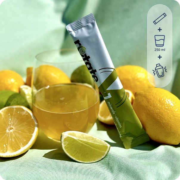 Rotate V1 Lemon Lime Stick neben einem Glas des zubereiteten Zitronen-Limetten-Getränks, umgeben von frischen Zitronen und Limettenscheiben auf mintgrünem Stoff
