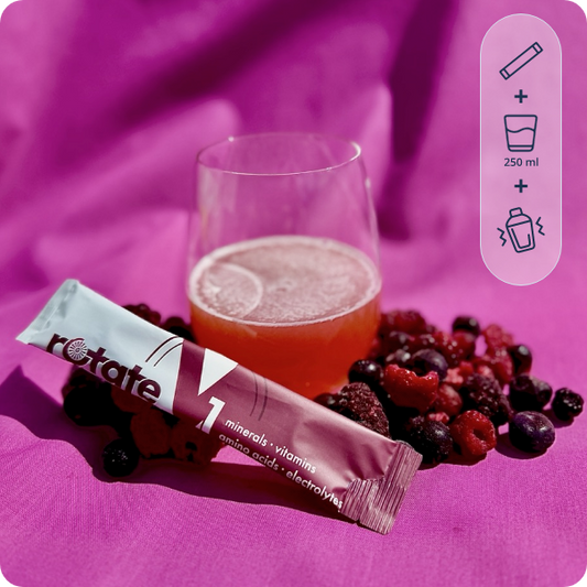 Rotate V1 Forest Fruit Stick neben einem Glas des zubereiteten Beerengetränks, umgeben von frischen Brombeeren und Himbeeren auf rosa Stoff