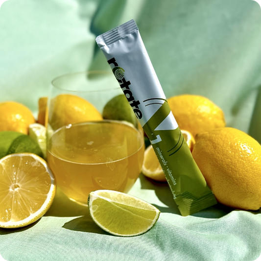 Rotate V1 Lemon Lime Stick neben einem Glas des zubereiteten Zitronen-Limetten-Getränks, umgeben von frischen Zitronen und Limetten auf mintgrünem Stoff