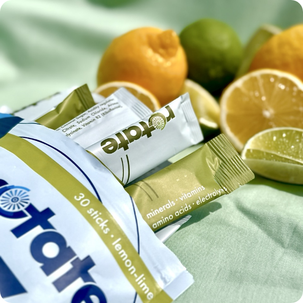 Rotate V1 Lemon Lime Verpackung mit 30 Sticks, mehrere Sticks angelehnt, auf mintgrünem Stoff mit Zitronen- und Limettenhälften