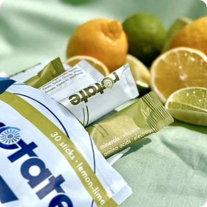 Rotate V1 Lemon Lime Verpackung mit 30 Sticks, mehrere Sticks angelehnt, auf mintgrünem Stoff mit Zitronen- und Limettenhälften